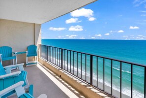 Ein Balkon mit türkisfarbenen Stühlen mit Blick auf einen Strand und klares, blaues Wasser, begrenzt durch ein Metallgeländer.