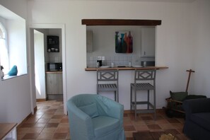 Espaço compacto com cozinha americana, dois bancos altos, uma poltrona azul e uma cadeira de madeira.