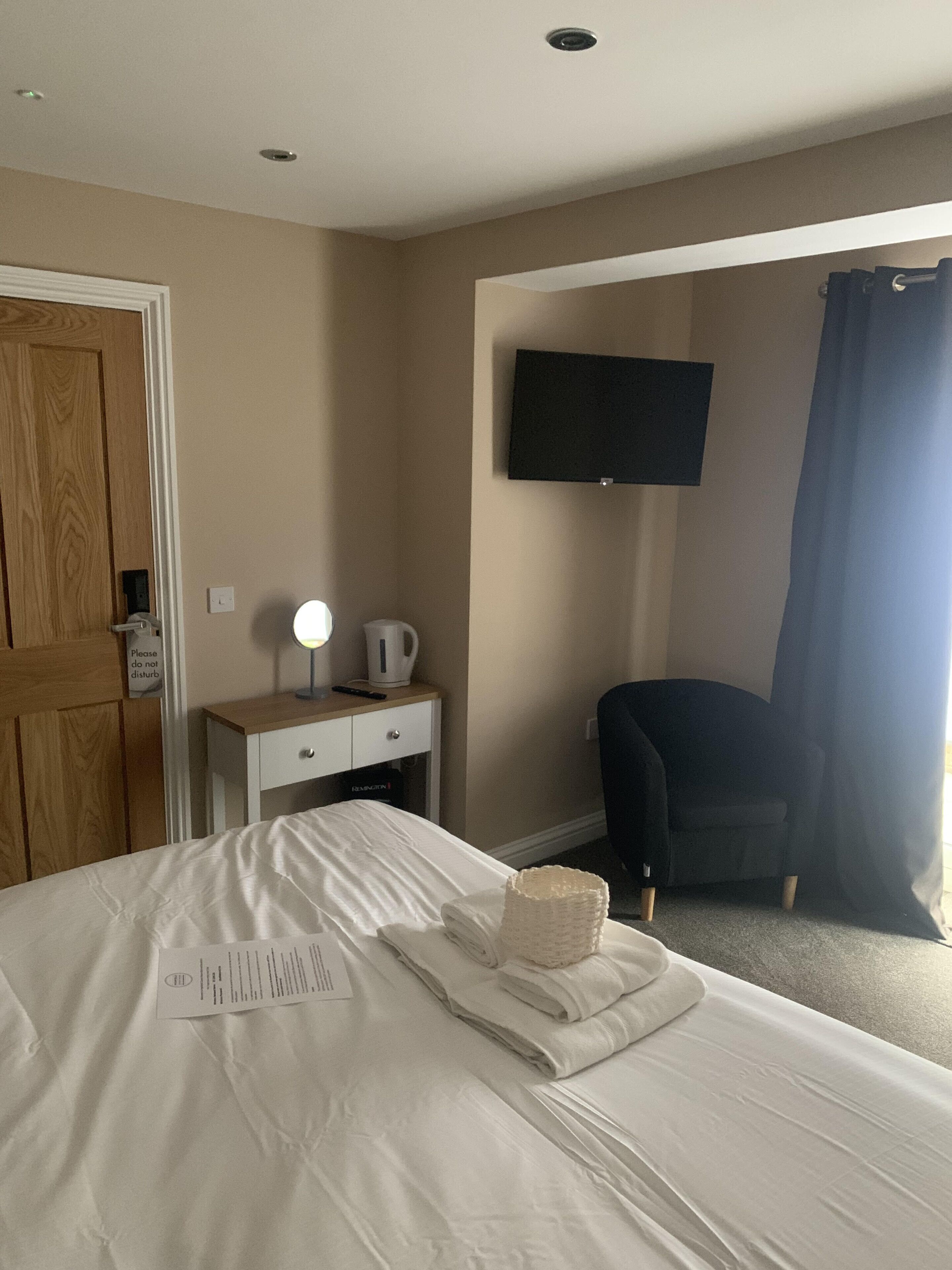 A hotel room with a bed, a desk with a lamp, a chair, a TV mounted on the wall, and a door.