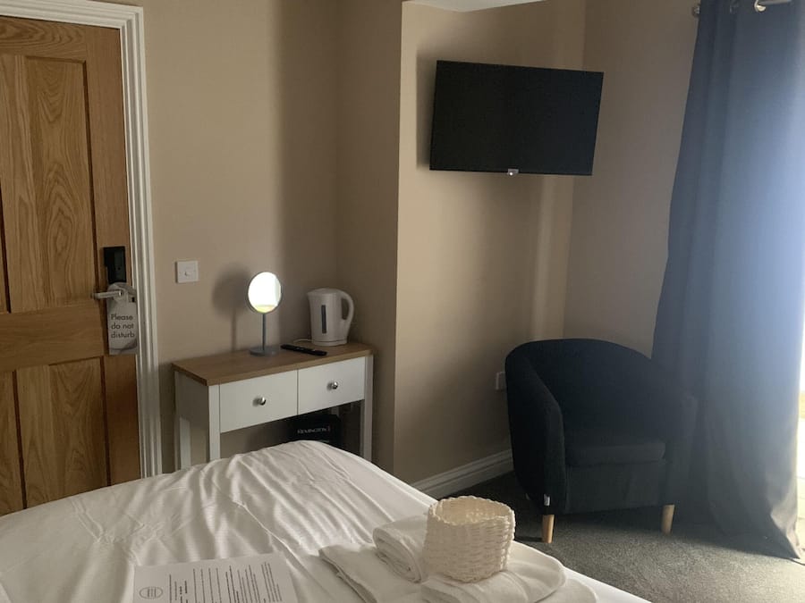 A hotel room with a bed, a desk with a lamp, a chair, a TV mounted on the wall, and a door.