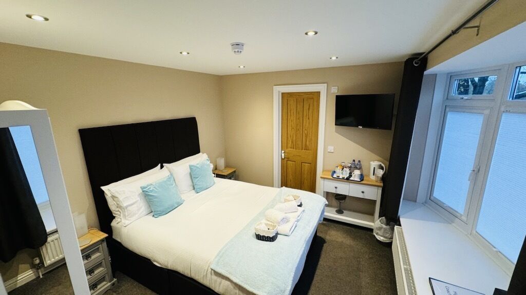 A hotel room with a bed, a bedside table with a lamp, a TV mounted on the wall, and a window.