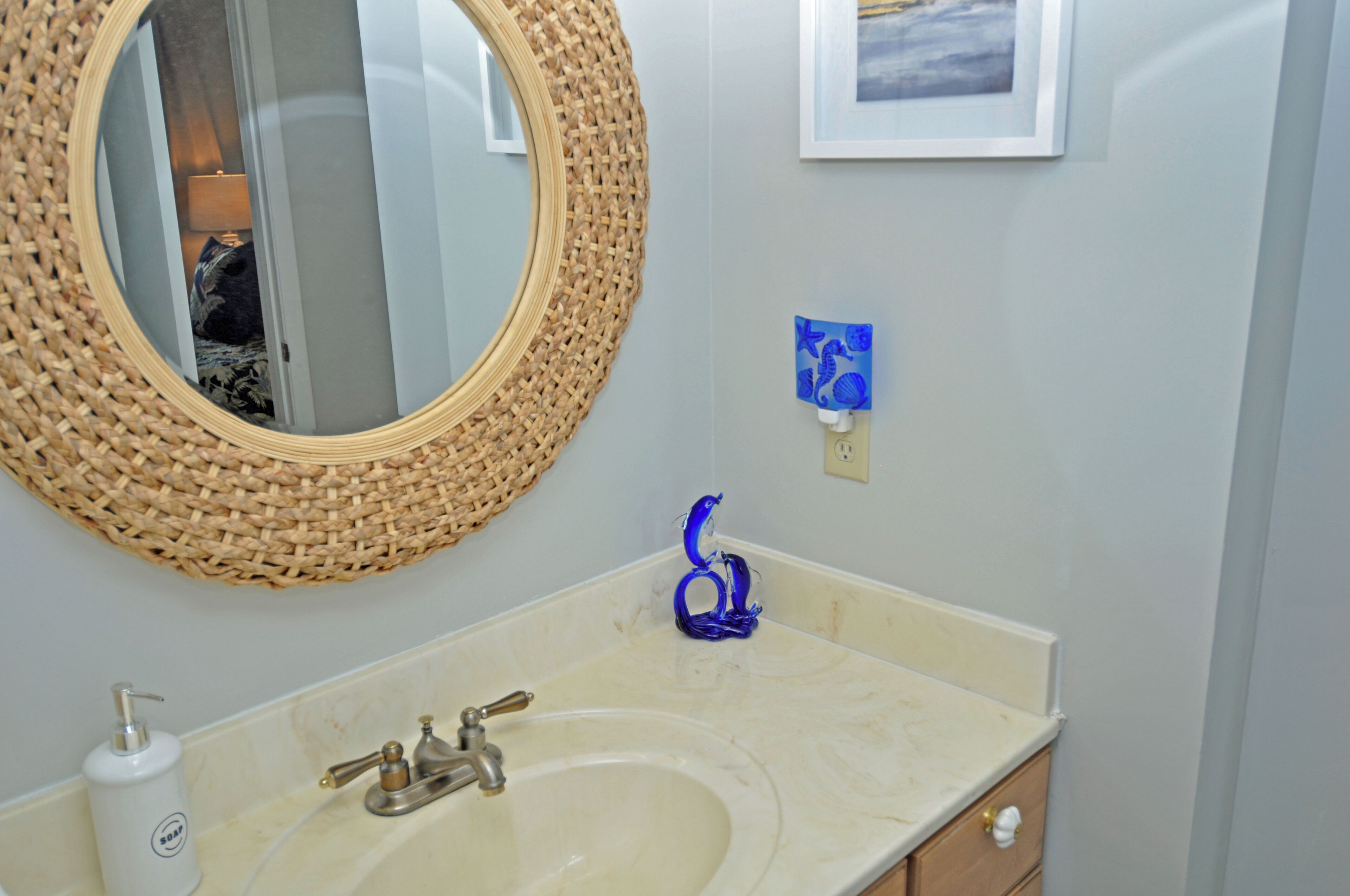 A bathroom with a round woven mirror, a sink with a single faucet, a small framed picture on the wall, and a soap dispenser on the counter.
