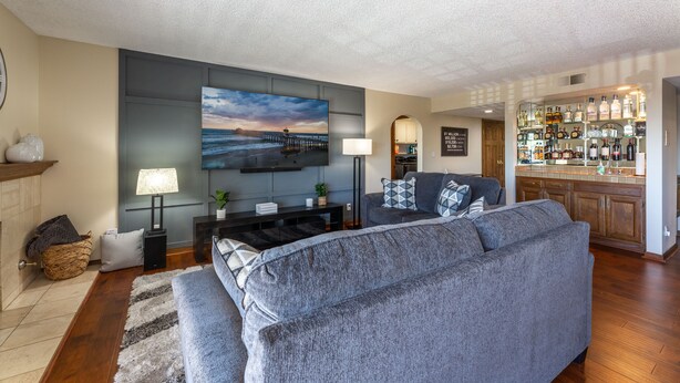 Una sala de estar con un sofá grande de color gris, un televisor de pantalla plana montado en la pared, una zona de bar con estantes llenos de botellas de licores, y piso de madera.