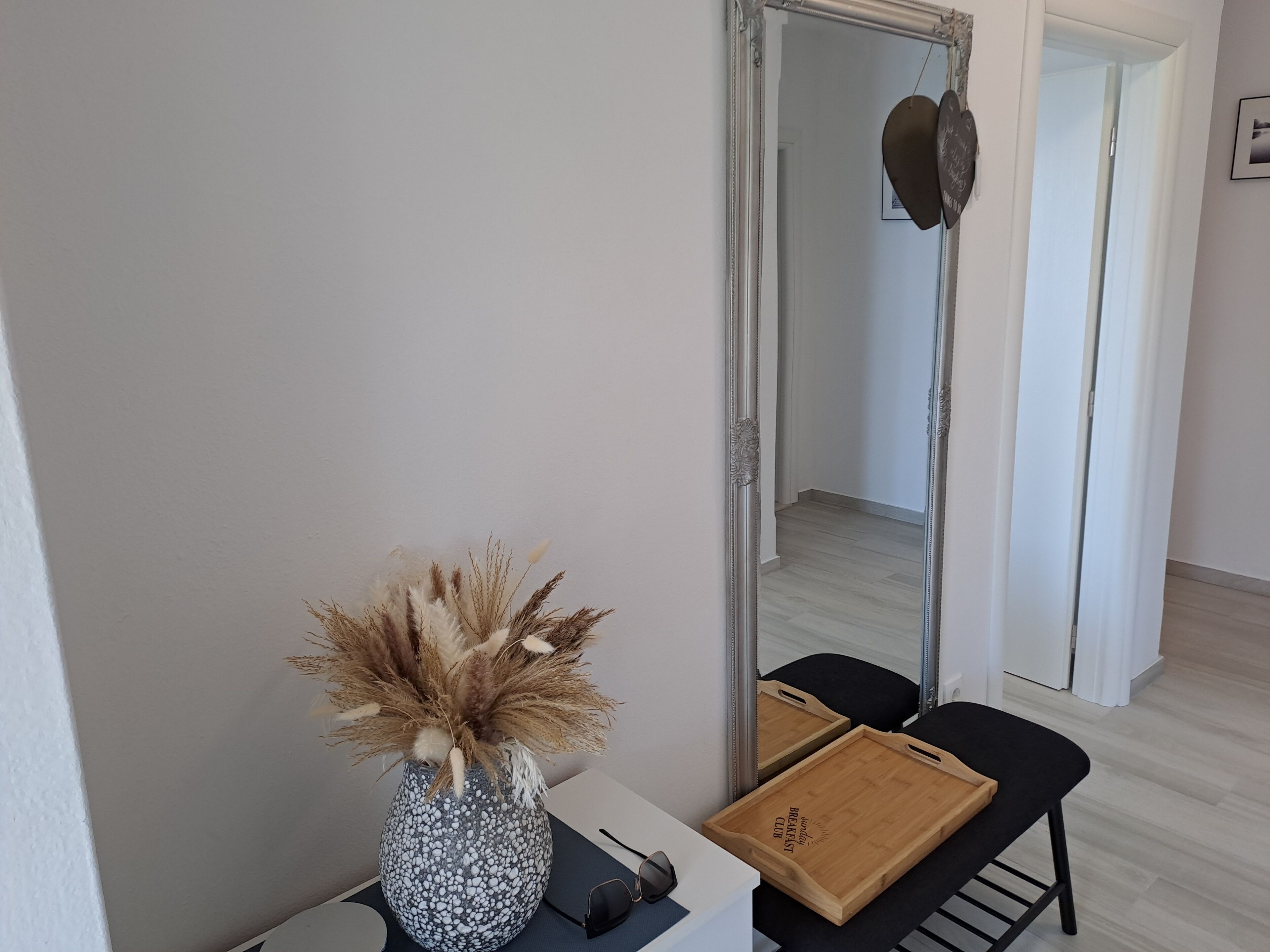 Een kamer met een spiegel, een vaas met gedroogde planten, een dienblad met ontbijtartikelen en een zwarte stoel.