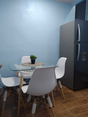 A compact kitchen with a glass dining table, white chairs, a potted plant, and a modern refrigerator.