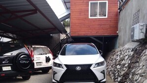 A white Lexus SUV parked in a driveway with a stone wall and a red building in the background.