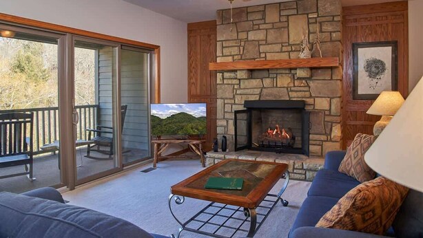 Ein gemütliches Wohnzimmer mit Kamin aus Stein, Deckenventilator und großem Fenster mit Blick auf eine Terrasse.