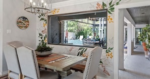 Uma área de refeições com mesa de madeira, cadeiras estofadas brancas, uma janela ampla e um detalhe decorativo na parede.