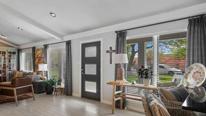 A spacious living room with a sofa, a coffee table, and a side table. There is a door with a cross on it, and a window with blinds.