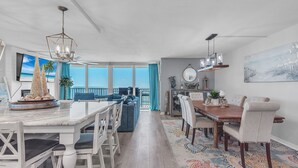 A spacious dining area with a large wooden table and white chairs, a ceiling fan, and a view of the ocean through large windows. - ☀Beachfront Condo🌴Stunning Updates👙Heated Pool🐬Trillium 3A🏖️Beach Access (Madeira Beach)