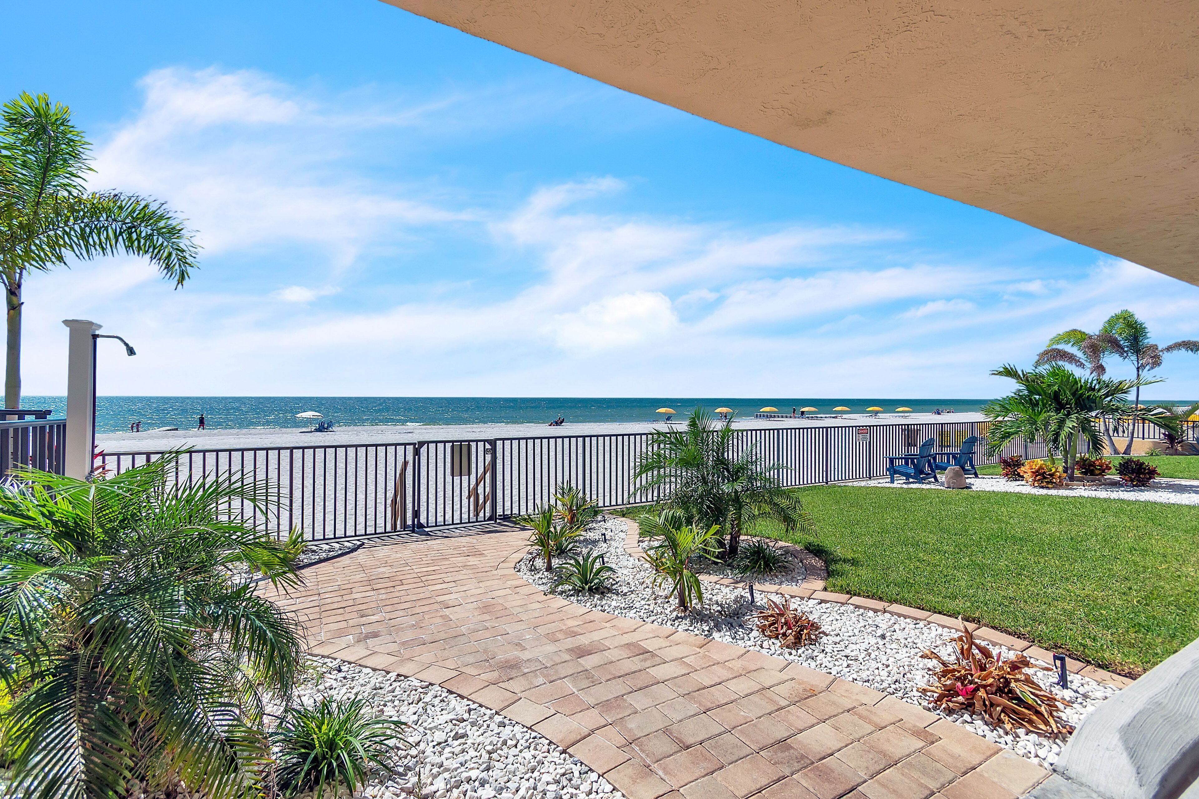 A paved patio with a view of the beach, palm trees, and a fence.