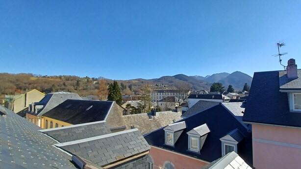 Il s’agit d’un quartier résidentiel composé de plusieurs maisons, dont certaines ont des toits en pente et d’autres des toits plats. On aperçoit des montagnes à l’arrière-plan, sous un ciel bleu dégagé.