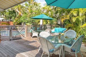 Un espace extérieur abrité, doté d’une table en verre entourée de chaises en osier, d’un parasol turquoise et d’une piscine en arrière-plan.