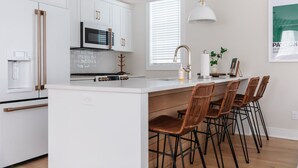 Une cuisine moderne avec des meubles blancs, un îlot central doté de tabourets de bar, un four encastré et un luminaire suspendu.