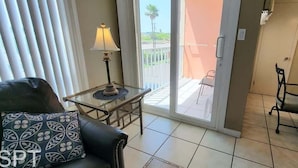 A living room with a sofa, a small table with a lamp, a balcony with a chair, and a dining area with chairs and a table.