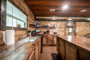 Une cuisine de style rustique, avec des éléments en bois, comprenant un îlot central, des placards en bois, un évier et une fenêtre équipée de stores.