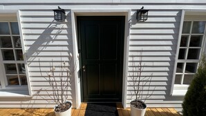 Un edificio blanco con una puerta principal oscura, dos plantas en macetas a cada lado y un felpudo negro.