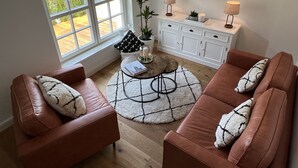 Una sala de estar con dos sofás de cuero marrón, una mesa redonda, un aparador blanco, un televisor montado en la pared y una ventana con persianas.