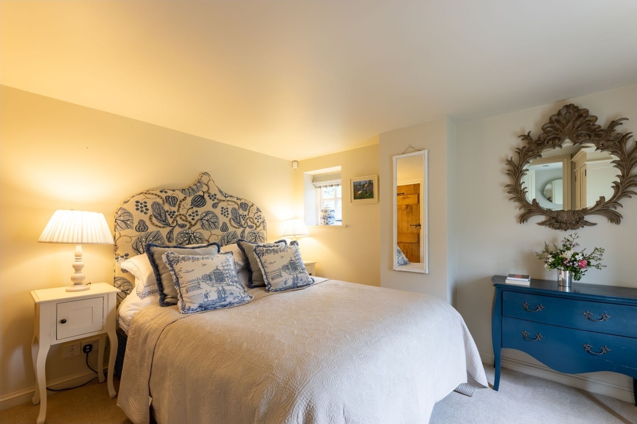 Une chambre à coucher avec un grand lit, une table de chevet avec une lampe, une commode avec des tiroirs, un miroir et un mur orné d’un motif décoratif.