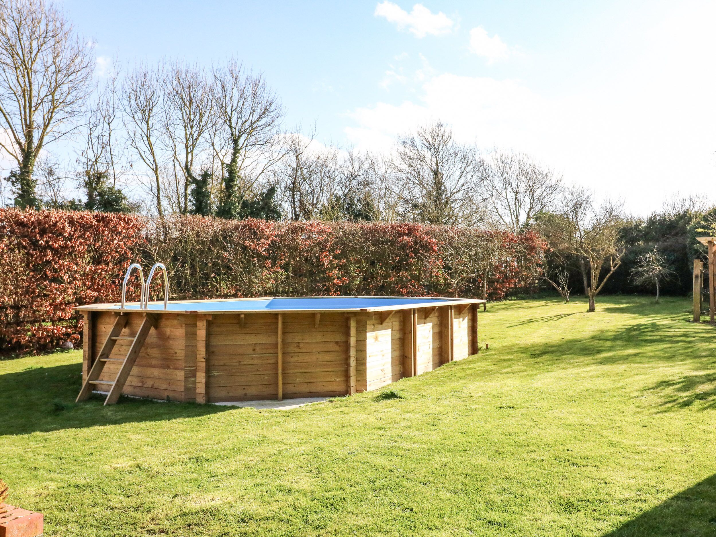 A wooden swimming pool with a ladder, surrounded by a well-maintained lawn and a hedge.
