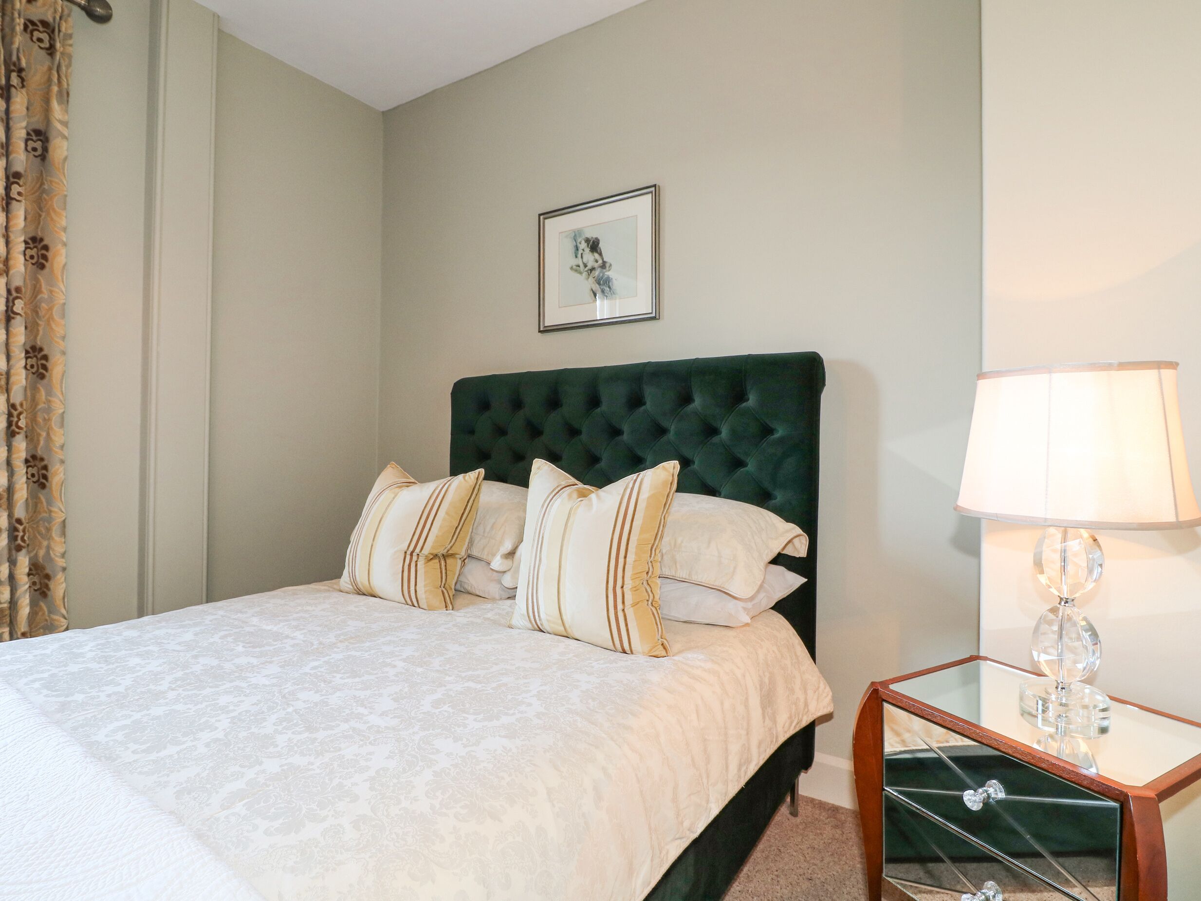 A neatly made bed with a green upholstered headboard, a bedside table with a lamp, and a framed picture on the wall.
