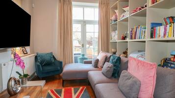 Un salón con un sofá gris, un sillón azul, una estantería llena de libros y un ventanal con cortinas.