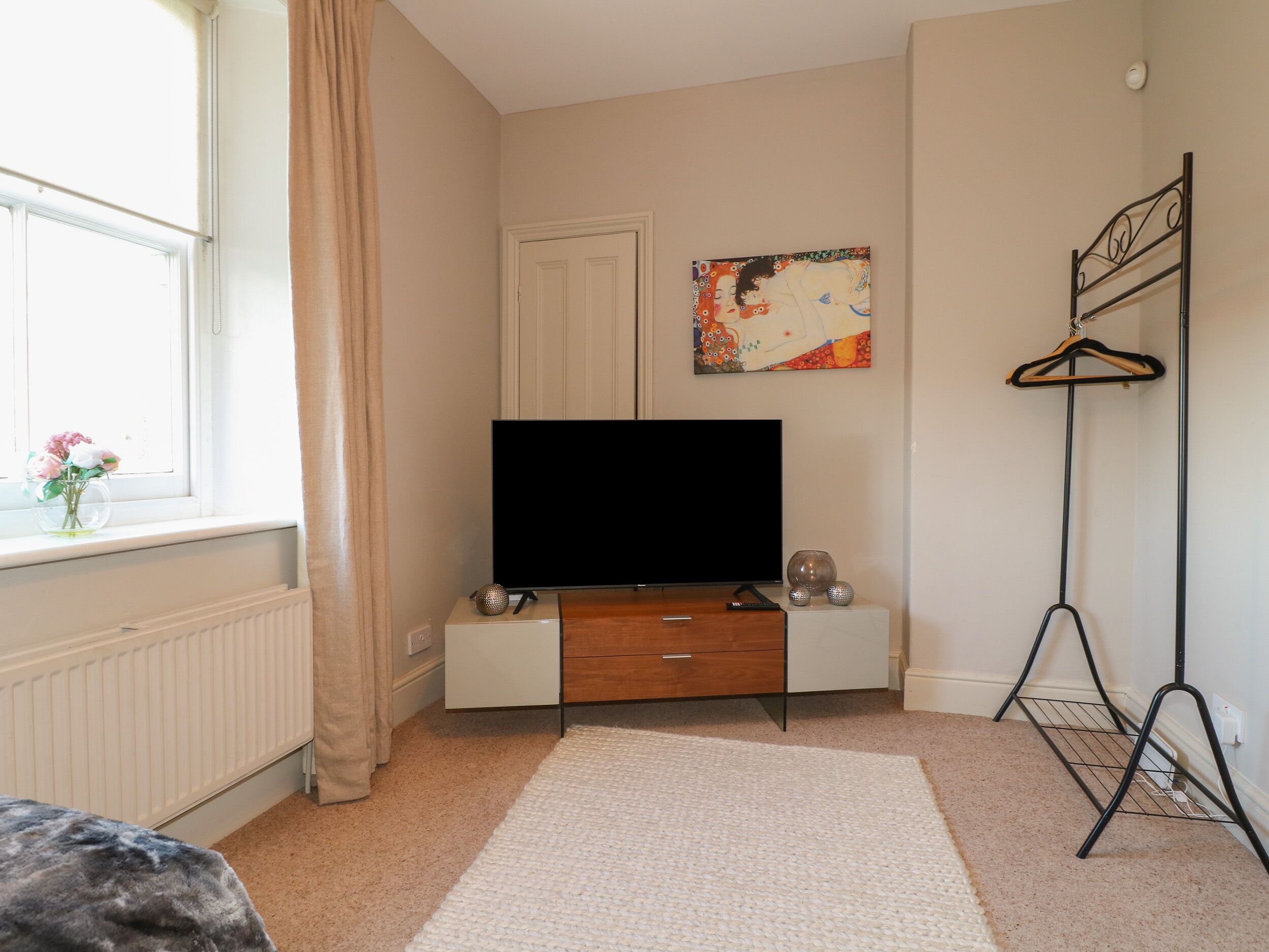 A bedroom with a television on a wooden stand, a radiator, a window with curtains, and a painting on the wall.