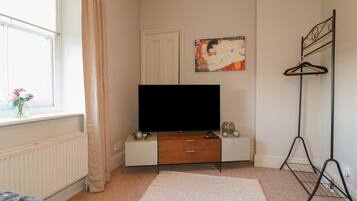 A bedroom with a television on a wooden stand, a radiator, a window with curtains, and a painting on the wall.