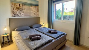 Une chambre à coucher avec un grand lit, un tableau représentant une plage, une table de chevet avec une lampe et une fenêtre avec des rideaux.