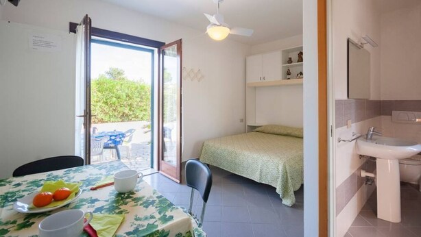 Une chambre avec un lit, une table sur laquelle se trouve une assiette de fruits, une chaise, un ventilateur de plafond et une salle de bain avec un lavabo.