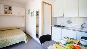 Une pièce de taille modeste comprenant un lit, une chaise, une table avec des fruits, un coin cuisine et une porte donnant sur une autre pièce.