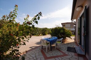 Eine Terrasse im Freien mit Tisch und Stühlen, umgeben von viel Grün und einem Gebäude.