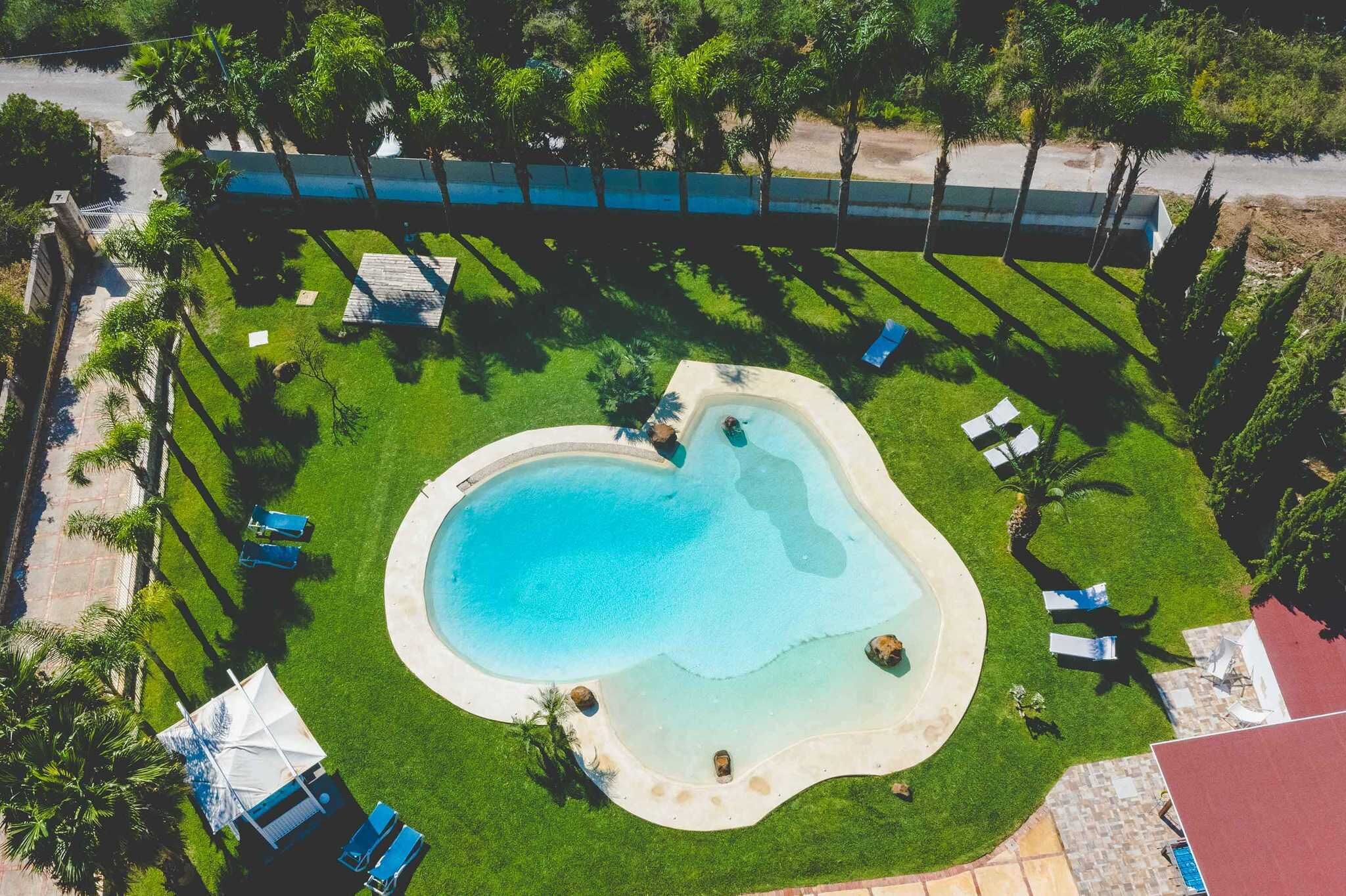 Ein Luftbild eines Schwimmbeckens, umgeben von Gras, Palmen und Liegestühlen.