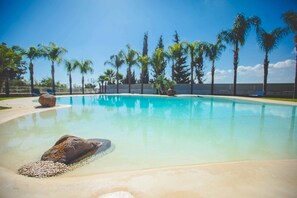A clear swimming pool with a rock in the center, surrounded by palm trees and a clear sky. - Flora apartment, in Casale Santuzza, with swimming pool (SIRACUSA)