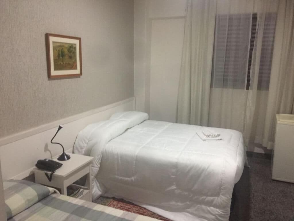 A neatly arranged hotel room with a bed, a bedside table with a lamp, a framed picture on the wall, and a window with curtains.