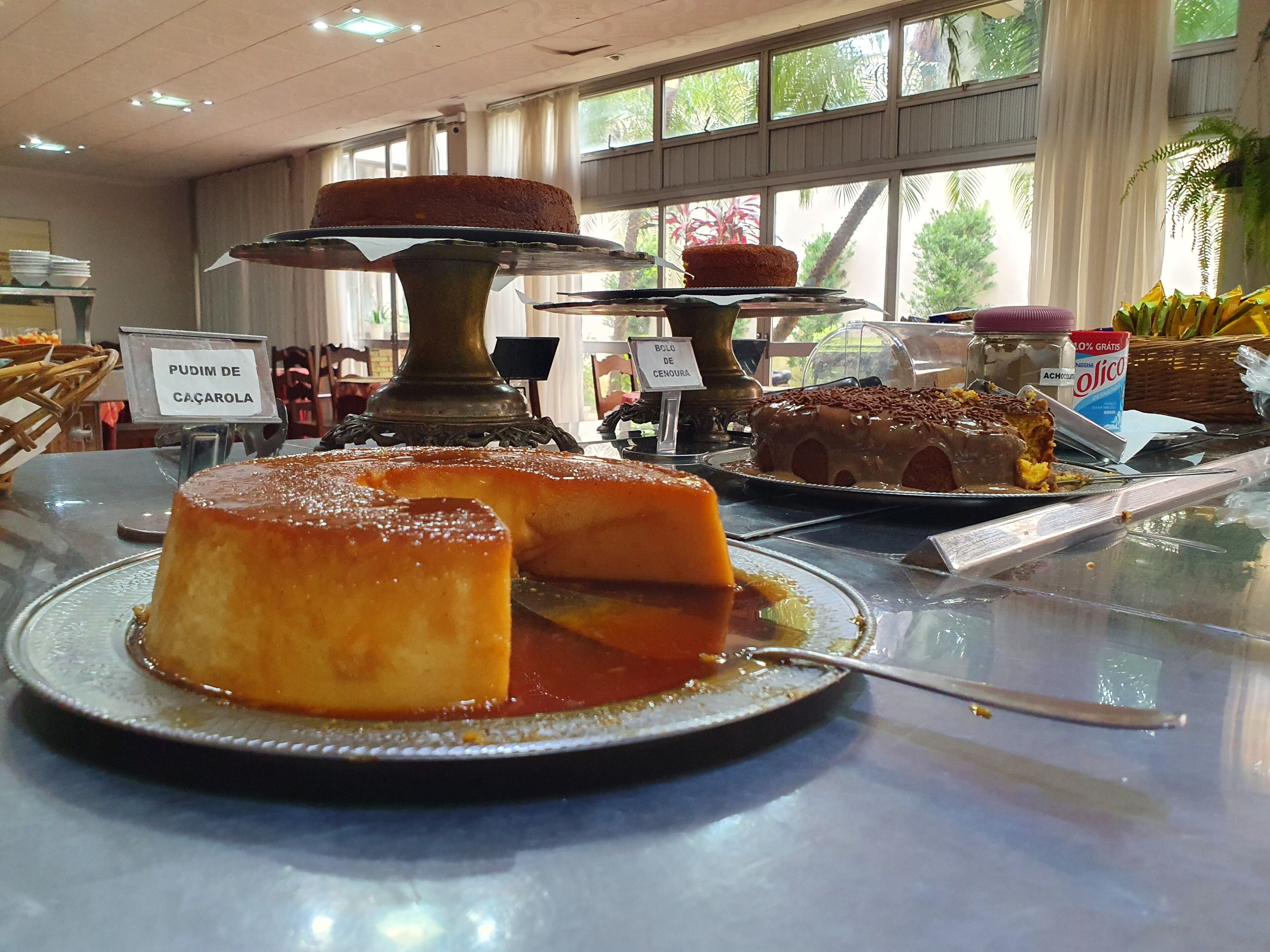 Una presentación de postres con diversos pasteles y pudines, incluyendo un pudín de caramelo y un pastel de chocolate, en una habitación bien iluminada con ventanas grandes.