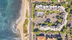 An aerial view of a coastal property with a beach, parking, and buildings with a pool.