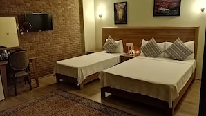 A hotel room with two single beds, a desk with a chair, a TV mounted on a brick wall, and two framed pictures on the wall. - Casa Callinos (Selçuk)