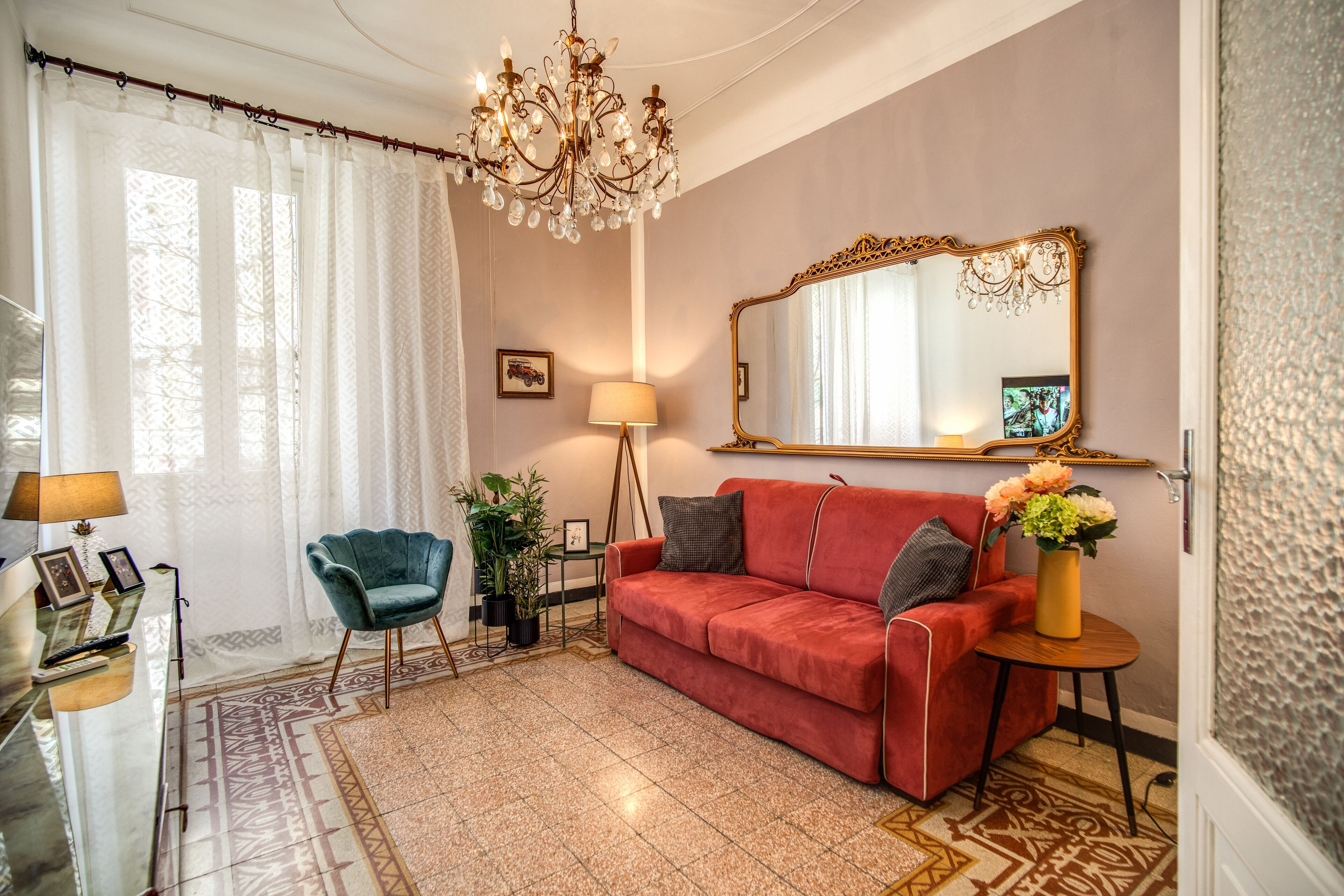 A living room with a red sofa, a blue armchair, a chandelier, a large mirror, and a small table with a vase of flowers.