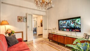Una sala de estar con un sofá rojo, un televisor montado en la pared mostrando varios programas, un mueble de madera y una alfombra estampada.