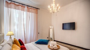 Habitación de hotel con cama, almohadas, televisor, candelabro y ventana con cortinas.