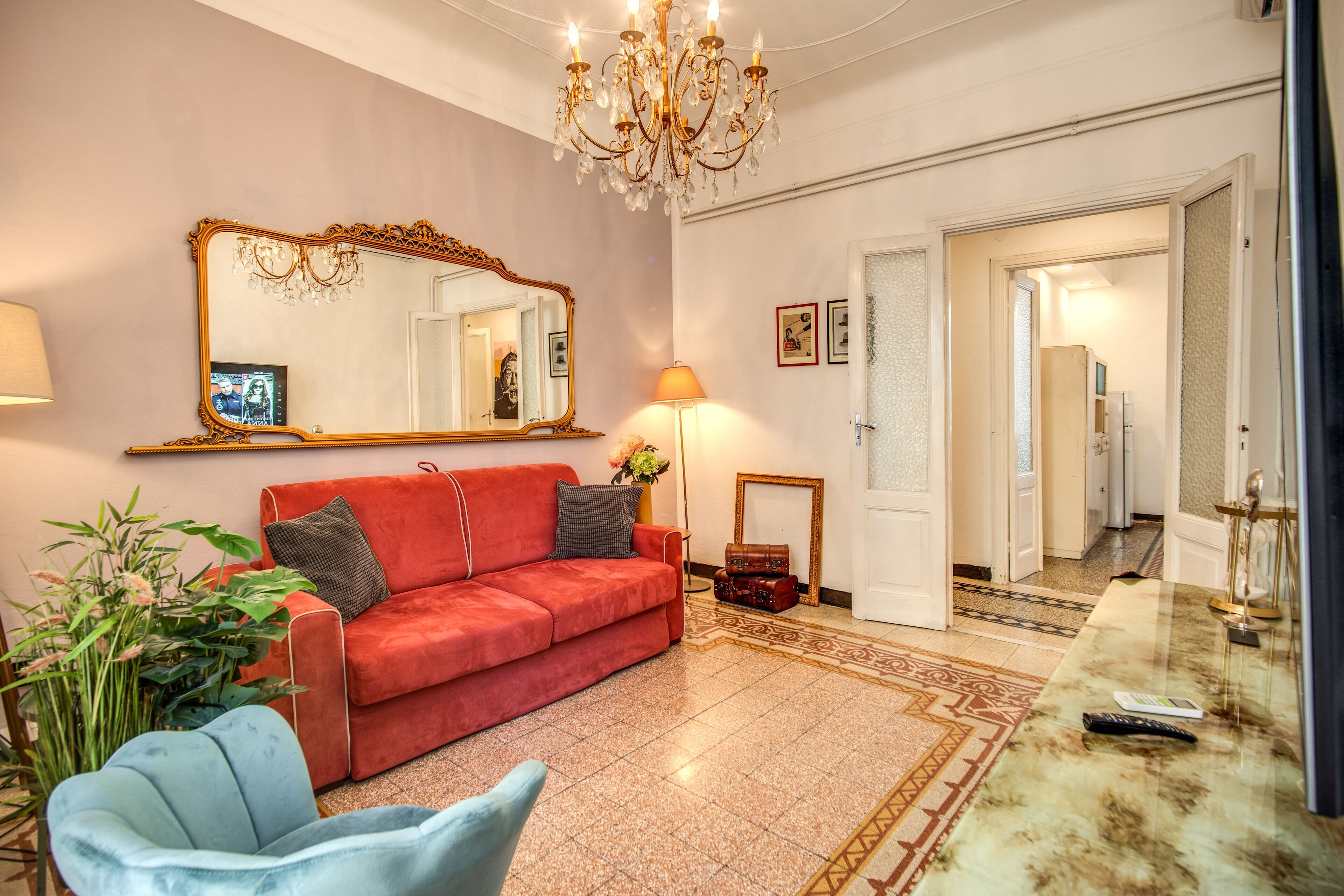 Un salon bien éclairé, avec un canapé rouge, un fauteuil bleu, un grand miroir, un lustre et une table basse en marbre.