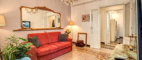Un salon bien éclairé, avec un canapé rouge, un fauteuil bleu, un grand miroir, un lustre et une table basse en marbre.