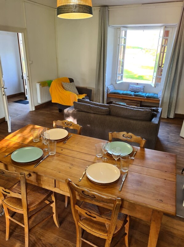 Uma sala de jantar com mesa de madeira posta para quatro pessoas, cercada por cadeiras de vime. Um sofá e um assento à janela com almofadas são visíveis ao fundo.