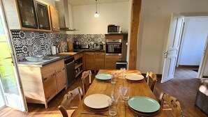 A wooden dining table set for four with plates and glasses, a kitchen with wooden cabinets and a microwave, and a ceiling with wooden beams and pendant lights.