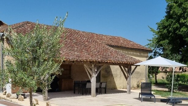 A swimming pool with a tiled deck, a covered patio area with outdoor furniture, and a potted tree.