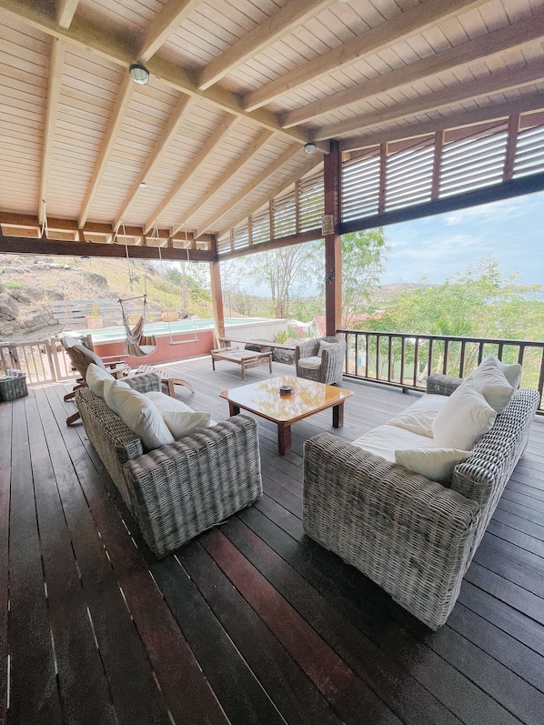 A covered outdoor seating area with wicker furniture and a wooden table, overlooking a scenic landscape.
