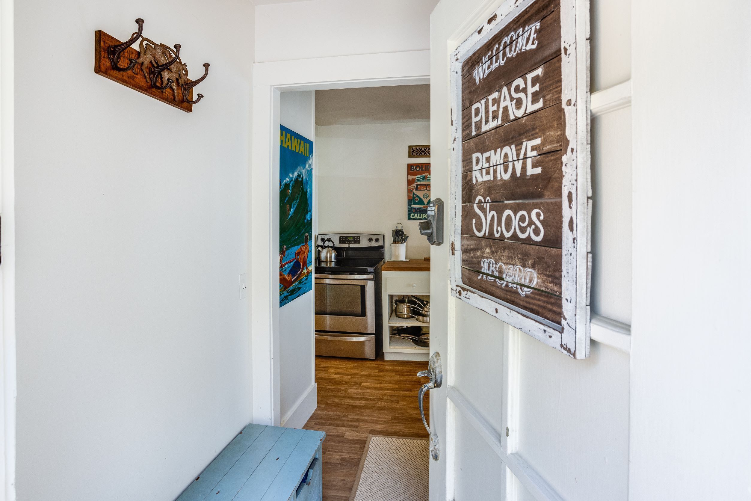 A hallway leading to a kitchen with a 'Please Remove Shoes' sign, a 'Welcome' sign, and a 'Hawaii' poster.