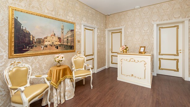 Una habitación con un cuadro grande enmarco, dos sillas ornamentadas, una mesita con un florero y un aparador blanco con detalles dorados.