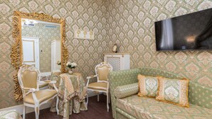 A room with a patterned wallpaper, a large ornate mirror, a sofa with patterned cushions, a small table with chairs, and a cabinet with a decorative top.